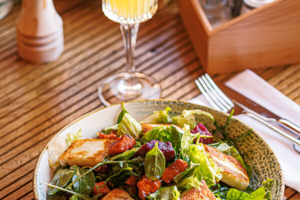 A bowl of leafy salad topped with grilled cheese squares and roasted vegetables, served with a bright orange drink garnished with an orange slice on a wooden table.