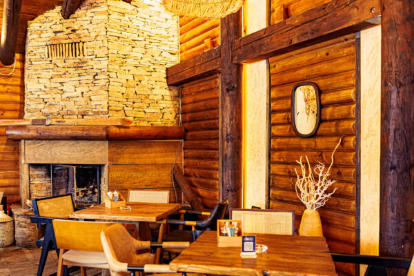 A lodge-style restaurant interior featuring a stacked stone fireplace, wood-paneled walls, and wooden tables with mixed upholstery.