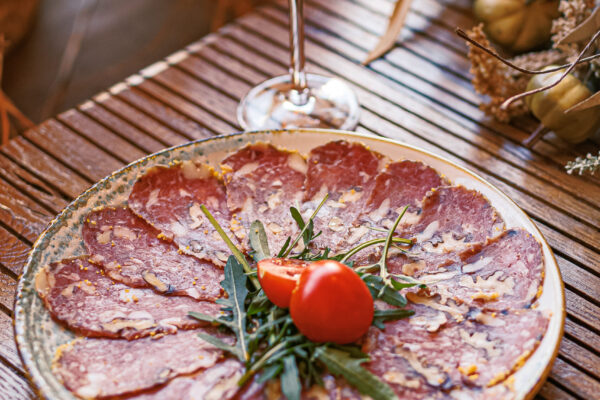 A plate of thinly sliced cured meat arranged in a circular pattern, garnished with a halved tomato and arugula, next to a tall glass of amber-colored liquor on a wooden table.