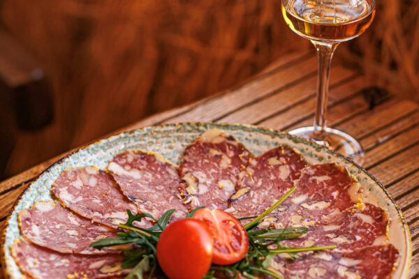 A circular arrangement of thinly sliced cured meat on a plate, garnished with a halved tomato and arugula, next to a glass of amber-colored liquor.