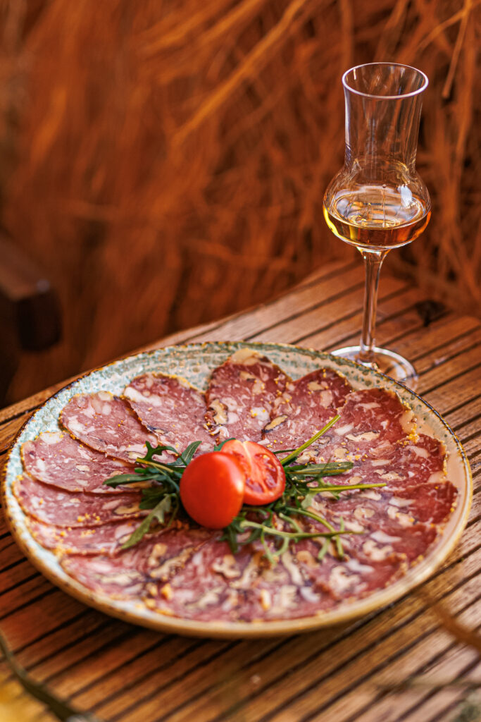 A circular arrangement of thinly sliced cured meat on a plate, garnished with a halved tomato and arugula, next to a glass of amber-colored liquor.