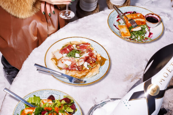 Two individuals wearing fur coats seated in the snow, toasting with champagne while plates of food and a bottle of Moët & Chandon rest on a makeshift snow table.