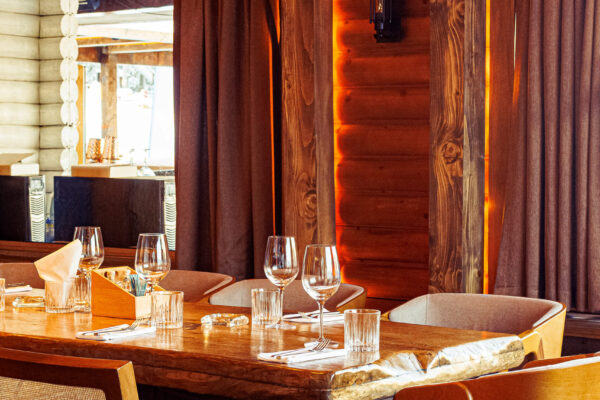A rustic restaurant interior featuring a wooden table set with glassware, surrounded by chairs, decorative gourds overhead, and warm amber lighting.