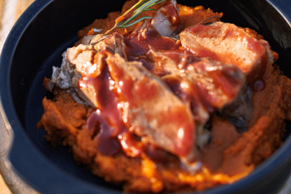 A black bowl filled with sliced meat on a bed of orange puree, drizzled with sauce and garnished with a sprig of rosemary.