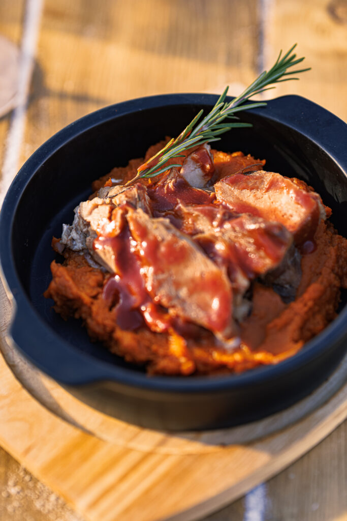 A black bowl filled with sliced meat on a bed of orange puree, drizzled with sauce and garnished with a sprig of rosemary.