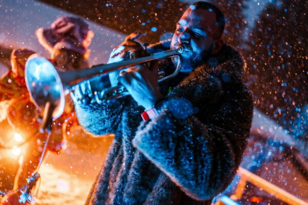 A musician wearing a fur coat plays a trumpet outdoors in falling snow, illuminated by colorful stage lights.