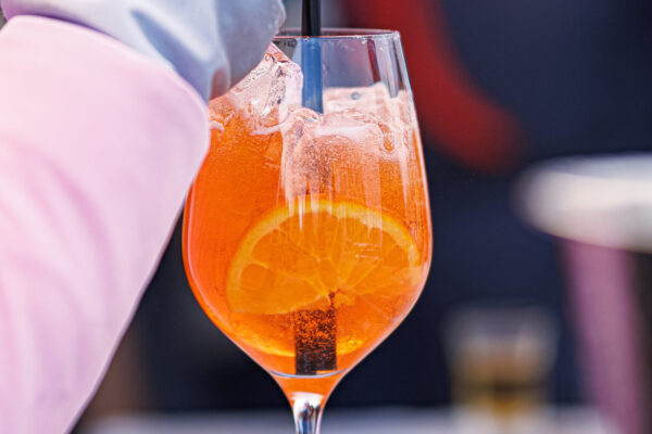 A close-up of an orange-colored cocktail in a tall glass with ice and an orange slice, being stirred by a person wearing a pink sleeve.