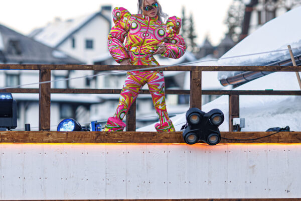 A performer wearing a brightly patterned outfit with exaggerated shoulders stands on a raised wooden deck, entertaining a crowd gathered below in a winter resort setting.