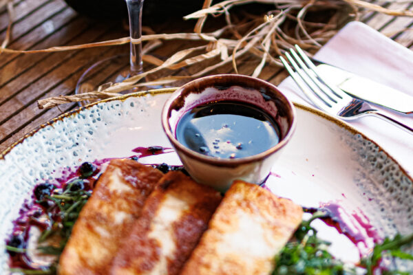 A white ceramic plate holding three seared cheese slices on a bed of greens, served with a dark dipping sauce, alongside a glass and bottle of red wine.