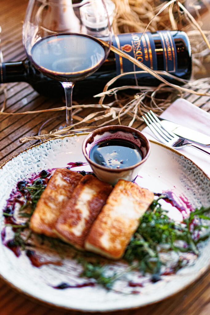A white ceramic plate holding three seared cheese slices on a bed of greens, served with a dark dipping sauce, alongside a glass and bottle of red wine.