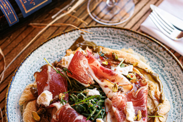 A round plate topped with thin slices of prosciutto, cheese, and greens on a crisp base, accompanied by a glass of red wine and a wine bottle on a rustic table.