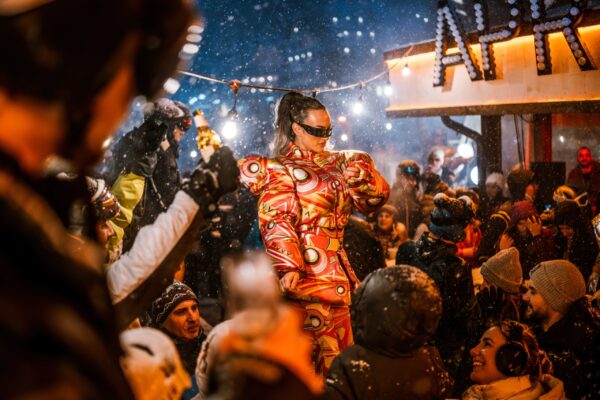 A performer wearing a brightly patterned snowsuit stands amid a crowd in falling snow, with an illuminated “APRES” sign in the background.