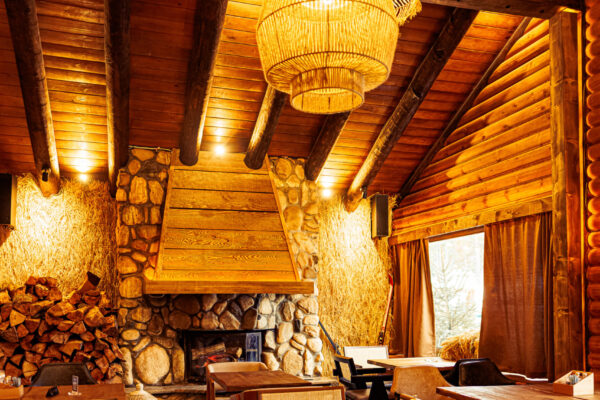 A high-ceilinged, lodge-style restaurant with a stone fireplace, stacked firewood, wooden tables, and large woven pendant lights.