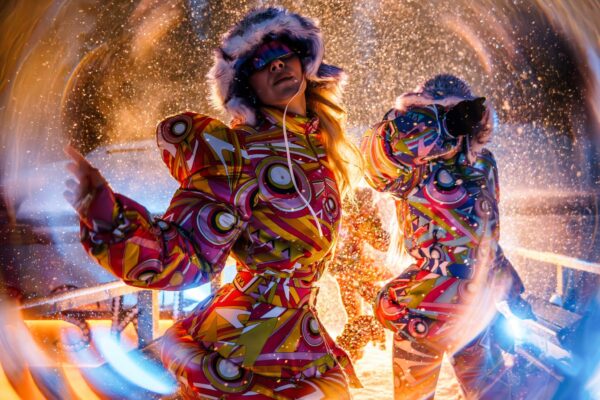 Two performers in matching, vibrant snowsuits with fur-trimmed hoods, posed under swirling lights and falling snow, creating a dynamic, tunnel-like effect around them.
