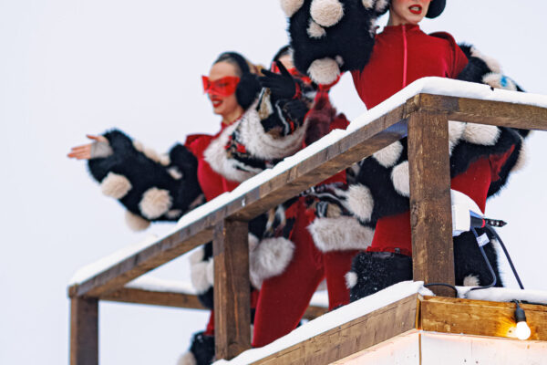 Two performers wearing matching red jumpsuits with black-and-white fur accents on their arms, standing on a raised platform and waving to a crowd in a snowy setting.