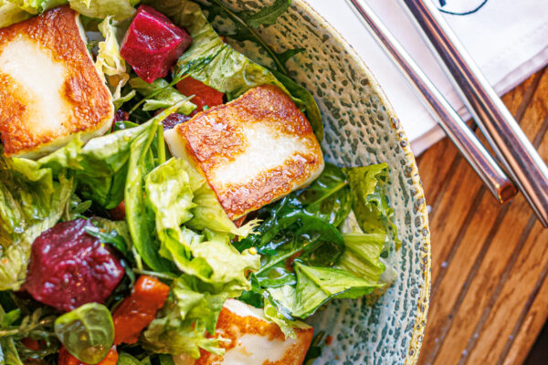 A bowl of mixed greens topped with golden-brown cheese squares and beet chunks, placed on a wooden table beside cutlery and a napkin marked with a stylized “A.”