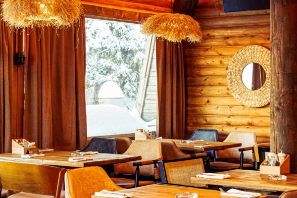 A cozy, wood-paneled dining area featuring straw-like hanging light fixtures, wooden tables, and a large window revealing a snowy view.