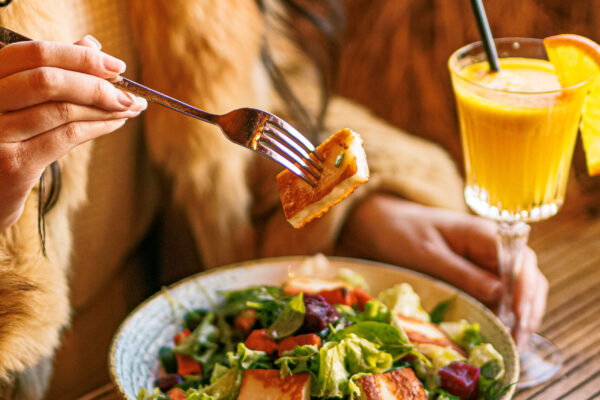 A woman wearing a fur-trimmed garment holds a fork with a piece of grilled cheese above a mixed salad, with a bright orange drink nearby.