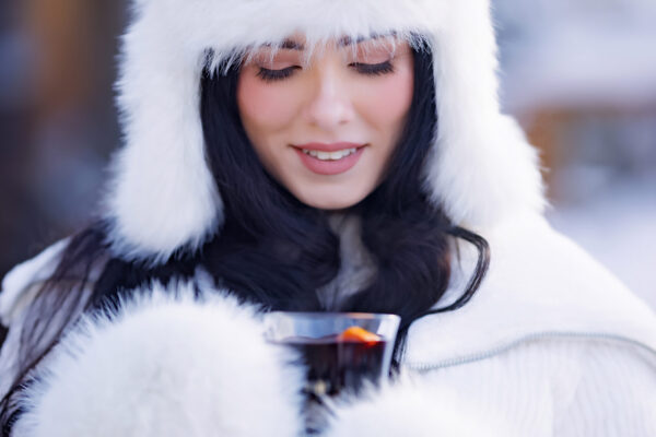 A woman wearing a white fur hat and coat, holding a steaming dark mug with an orange slice in a snowy setting.