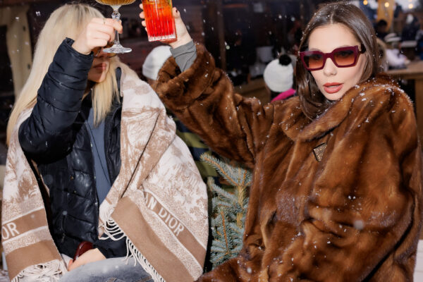 Two women in fashionable winter attire raising cocktails in front of an illuminated “APRES SKI” sign at a snowy outdoor bar.