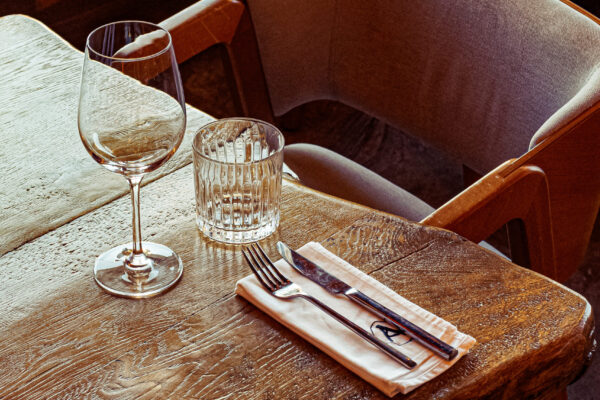 A rustic wooden table set with a wine glass, water glass, and neatly folded napkin holding utensils, next to a padded wooden chair.